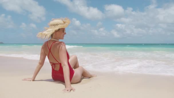 ビーチで日光浴を楽しむストロー帽子と赤い水着の美しい少女 バックビューの若い日焼けした女性は アゾールブルーの海の背景で風を楽しんでいます キャリーフリーの女性は笑顔で ハワイに頭を投げる — ストック動画