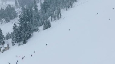 Kış tatili gezisi. Kış sporları arka planı. Kar fırtınasında tepelerden kayakçıların üstünde yavaş çekim yapan drone. Stevens Pass Washington 'daki kayak merkezindeki sinematik kış dağ ormanı.