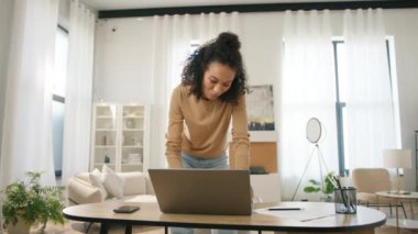 Dizüstü bilgisayarda çalışan melez bir kadın. Evde uzaktan çalışan pozitif renkli bir kadın. Modern oturma odasındaki çalışma güvertesinde hareketli arka planda duran neşeli, çoklu etnik girişimci.