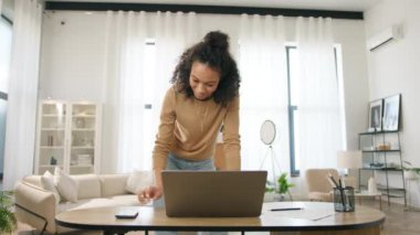 Evde uzaktan çalışan, renkli bir kadının telefonuna cevap veren biri. Neşeli meşgul girişimci modern oturma odasındaki çalışma güvertesinde duruyor. Dizüstü bilgisayarda çalışan gülümseyen melez kadın.