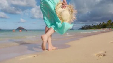 Hawaii, Lanikai sahilinde yaz tatilindeki kadın turist. Güzel sallanan pareolu kadın, hasır şapkalı. Yavaş çekim kadın ayakları kumsalda yalın ayak yürüyor altın gün batımı ışığında kumsalda ayak izleri bırakıyor