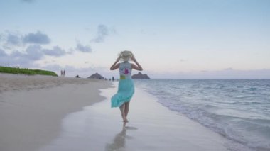 Mavi plaj elbiseli ve hasır şapkalı mutlu gezgin sinematik doğanın tadını çıkarıyor. Hawaii Adası 'ndaki tropik cennet turizm arka planının kopyası. Gün batımından sonra egzotik sahilde yürüyen genç kadını izleyen kamera 4K