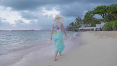 Gün batımından sonra egzotik sahilde yürüyen genç bir kadını takip eden kamera. Mavi plaj elbiseli ve hasır şapkalı turistler sinematik doğanın tadını çıkarıyorlar. Hawaii adasındaki tropik cennete seyahat et. Kopya alanı arka planı