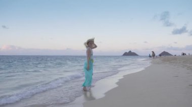 Mavi sahildeki mutlu gezgin hasır şapka takıyor ve Lanikai sahilinde Na Mokulua adalarıyla sinemanın tadını çıkarıyor. Tropik cennet Hawaii adası. Gün batımından sonra egzotik sahilde yürüyen genç bir kadın.