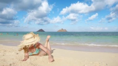 Lanikai plajı Mokulua adaları manzaralı güneşlenen kadın gezgin. Fırfırlı hasır şapkalı gülümseyen turist Oahu sahilinde altın kumda kalp çiziyor. Kumsalda bronzlaşan genç bir kadın. Aşk ve mutluluk kavramı