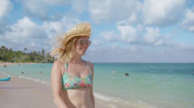 Kaygısız Amerikalı kadının mutlu yüzü Oahu arka planında deniz mavisi bir okyanusun kenarında yürüyor. Hasır şapkalı güzel kız Hawaii sahilinde güneşleniyor. Altın güneş gözlüklü genç kadın meltem 4K 'nin tadını çıkarıyor.