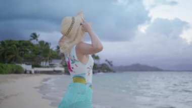 Fırfırlı hasır şapkalı neşeli bayan okyanus tropik plajında dikiliyor. Mutlu turist, Lanikai Sahili 'nde bulutlu bir esintinin keyfini çıkarıyor. Arka planda yeşil palmiye bahçesi var. 4K