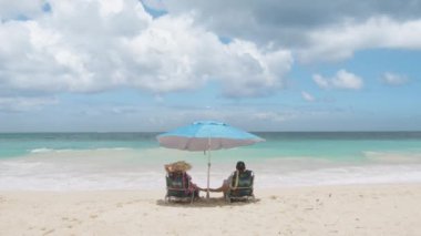 Açık havada dinlenen zengin turistler plaj sandalyelerinde mavi plaj şemsiyesiyle kaplanmış bir şekilde oturuyorlar. Birbirine aşık erkek ve kadının el ele tutuşup okyanusun tadını çıkarışının arka görüntüsü. Yaz tatilinde bronzlaşan gezginler
