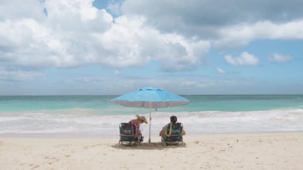 游客们坐在沙滩上的椅子上 遮掩着蓝色的沙滩伞 在户外放松身心 回顾男女坠入爱河的情景 享受蜜月之旅的海洋 游客在暑假晒日光浴4K — 图库视频影像