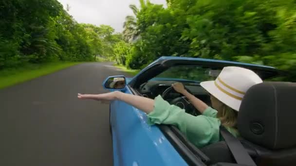 スタイリッシュな若い女性ドライバーの外側の青いコンバーチブルカーからの眺め バックビュー幸せな旅行者女性手を振り 風光明媚な緑豊かな熱帯の島で高速運転しながら空気をキャッチ バケーションの背景 — ストック動画