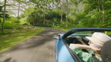 Hawaii 'li turist güneş ışınlarına uzanıyor. Mavi üstü açık arabadaki mutlu kız yaz gününün tadını çıkarıyor ve rüzgarı eliyle yakalıyor. Tarz sahibi kadın dağ manzaralı tropikal adalardan geçiyor.