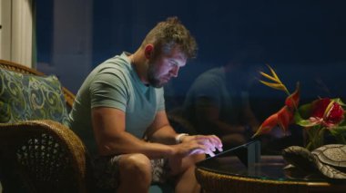 Yetişkin erkek, gece geç saatlerde tablet ekranda veya sosyal medya blogu içeriğinde yazıyor. Odaklanmış adam, parlak ekran teknik cihaza bakıyor. İş adamı uzaktan tropikal tatilde çalışıyor.
