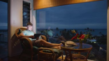 Okyanus manzaralı büyük bir duvar penceresinde oturan bir adam, geceleri kablosuz ağ kullanarak tablet ekrana bakıyor. Seyahat rotasını internetten keşfeden adam. Hawaii adası tarzı odasındaki tablette erkek arama içeriği