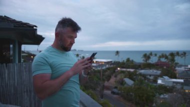 Olgun yakışıklı adam bulutlu bir akşamda tropikal ada manzaralı balkonda dikiliyor. Atletik adam akıllı telefon ekranına bakıyor. Sakallı işadamı Lancer 4K e-postalarına uzaktan cevap veriyor.