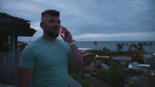 有胡子的生意人是自由的长矛手 工作着远程接听电话 在阴天 一个健康的男人站在有热带岛屿景色的阳台上 在外面用智能手机说话的有运动精神的微笑的家伙 — 图库视频影像