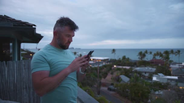 在阴云密布的夜晚 站在有热带岛屿景色的阳台上的英俊男子 一个运动员在看智能手机屏幕 有胡子的商人免费电话接线员 远程回复4K电子邮件 — 图库视频影像