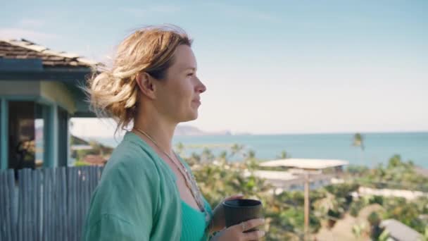 リラックスしたリネンシャツの若い女性は 紅茶のカップでバルコニーで ハワイ島で熱帯の休暇を楽しんでいるエレガントな女性 レジャーライフスタイル 笑顔の女性はビーチヴィラからの海の景色を見下ろす — ストック動画