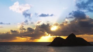 Parlak altın güneş ışınları okyanusun üzerindeki mavi bulutların arasından parlıyor. Hawaii adalarında dramatik doğa. Oahu macera gezisi. Na Mokulua adalarının üzerinde, Oahu adasının rüzgarlı sahillerinde nefes kesici bir gün doğumu.