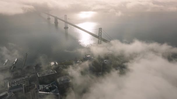 サンフランシスコ湾のベイブリッジの上のシネマティックミスト 輝く霧の下の黄金の日差しにある風光明媚な現代都市の景色 日の出にサンフランシスコのダウンタウンをカバーする黄金の雲のエピックドローン飛行 — ストック動画