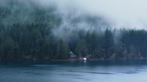 高山湖上的空中戏剧化 孤独的房子在河岸的常绿茂密的森林里 被电影般的雾覆盖着 在多愁善感的天气里 云彩飘浮在明镜蓝湖边的松树上 — 图库视频影像