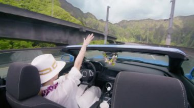 H3 otobanından geçen mavi üstü açık arabalı mutlu turist. Cabriolet 'in açık arabasından görülen manzara Hawaii manzarası. Ley çiçekli zarif kadın arabadan elini sallayarak indi. Kadın Oahu Adası 4K 'dan geçiyor.
