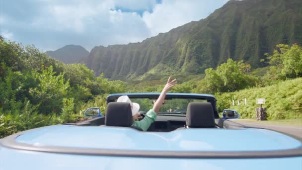 近代的なブルーのコンバーチブルカーのバックビューは離れて運転します 雄大なジャングル山の景色を楽しむドライバーの女性 女性は夏休みにオアフ島を旅行する ハワイのブルーコンバーチブルカーでハッピーな観光客 — ストック動画