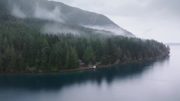 オリンピック国立公園のクレセント湖 シーニックワシントン自然4K アメリカ 風光明媚な湖での山林小屋の空中眺望 濃い霧で覆われた常緑の熱帯雨林に隠されたシネマティックな素朴な家 — ストック動画