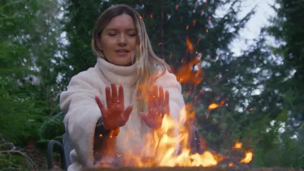 在绿色云杉森林背景的火坑外的满意的游客 一个微笑的女人在篝火中取暖的画像 快乐的女性享受周末远足时焚烧篝火的自然之旅 — 图库视频影像