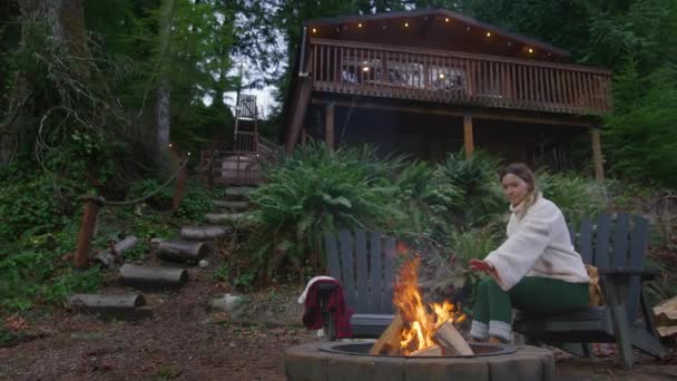 木製のキャビンで薪を燃やして手を温める女性 田舎の家の屋外でリラックスして緑の森で火のピットで燃える女性 週末の休暇 自然の目的地 アメリカ — ストック動画