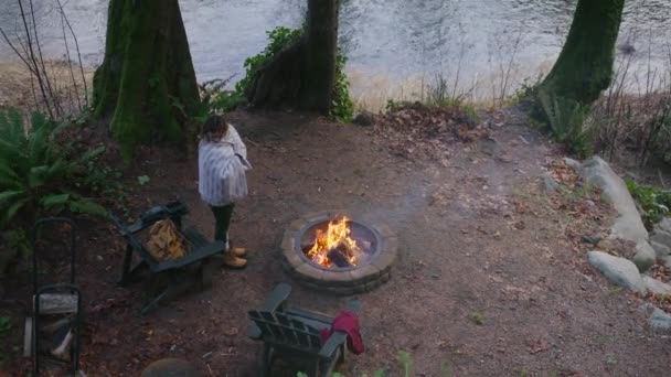 緑の川で燃える火を楽しむリラックスした女性 背景に緑のスプルースの森のファイヤーピットでキャンプ 週末の休暇中に暖まる居心地の良い毛布に包まれた女性のオーバーヘッドビュー — ストック動画