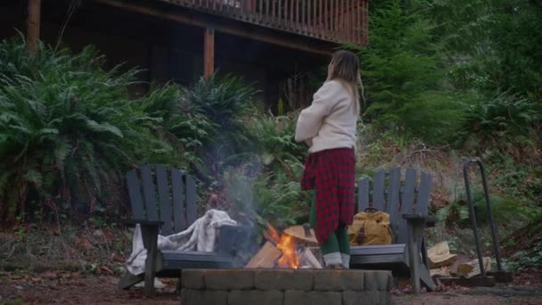 緑の森4Kで火事で田舎の家の屋外でリラックスした女性 遠隔地の自然の目的地アメリカでの週末の休暇 木製のキャビンで外の火のピットで木製のログを燃やしている若い女性 — ストック動画