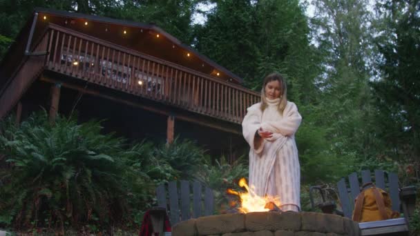 森の小屋で週末に薪を燃やして居心地の良い毛布で温まる女性 女性は背景に緑のスプルースの森で燃える火で手を温めます 燃える火を楽しむリラックスした女性 — ストック動画