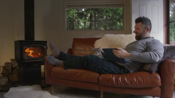 森林小屋でWifiを使用している男性は 木材の燃えるストーブで暖かくなります 人とテクノロジーを遠隔地に 快適な茶色の革ソファーでリラックスして モバイルタブレットでニュースを読むハンサムな男 — ストック動画