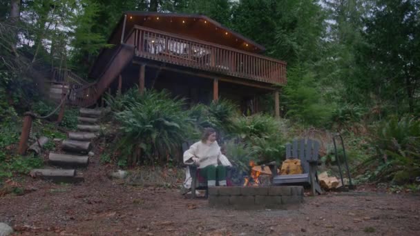 離れた自然の目的地での週末の休暇 現代の木製の小屋の外の火の穴で木製のログを燃やしている若い女性 緑の森で燃える火で田舎の家の屋外でリラックスする女性 — ストック動画