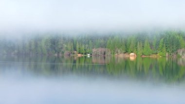 Dağ ormanlarında, alçak sis bulutu içinde rıhtım kenarındaki ahşap kulübeler. Sinema havası dağ gölü manzarası. Durgun su yüzeyine yansıyan ebedi yeşil ormanın huzurlu manzarası