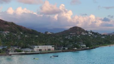 Yaz tatili geçmişi. 4K Oahu turizmi. Lanikai sahilinin yukarısındaki yeşil tepeye yuva yapmış tatil evleri. Şafak vakti tropikal manzara üzerinde hava manzarası. Güneş Hawaii adasının üzerinde bulutları aydınlattı