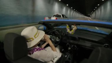 Oahu adasını keşfeden kadın sürücü. Güvenli sürüş konsepti 4K. Kadın turist dağ tünelinde hızlı bir şekilde üstü açık araba kullanıyor. Modern fütüristik tünelde beyaz şapkalı bir gezginin arka koltuk görüntüsü.