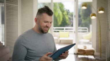 Tablet ekranına bakıp yüksek sesle gülen mutlu bir adam. Aşırı neşeli orta yaşlı erkek, tablet üzerinde eğlenceli komik video içeriği arıyor. İnsanlar modern teknolojiyi ev 4K 'de eğlence için kullanıyorlar.