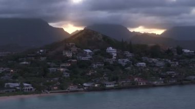 Lanikai sahilinin yukarısındaki tepedeki modern sahil kulübeleri. Şiddetli yağmur bulutlarıyla kaplı yüksek dağların ardında altın gün batımı görülüyor. Hawaii, Oahu Adası 'ndaki lüks okyanus paravan evleri. Gayrimenkul yatırımı