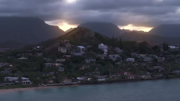 ランカイビーチの高い丘の上の近代的なビーチコテージ 豪雨の雲に覆われた高い山の後ろに見られる黄金の日没 ハワイのオアフ島にある高級オーシャンフロントハウス 不動産投資について — ストック動画