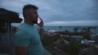 Bulutlu bir günde tropik ada manzaralı balkonda duran sakallı sağlıklı bir adam. Atletik gülen adam dışarıda akıllı telefon kullanarak konuşuyor. İşadamı serbest mızrakçı. Çağrıya cevap veriyor.
