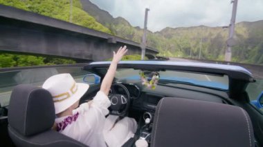 H3 otobanından geçen mavi üstü açık arabalı mutlu turist. Cabriolet 'in açık arabasından görülen manzara Hawaii manzarası. Ley çiçekli zarif kadın arabadan elini sallayarak indi. Kadın Oahu Adası 4K 'dan geçiyor.