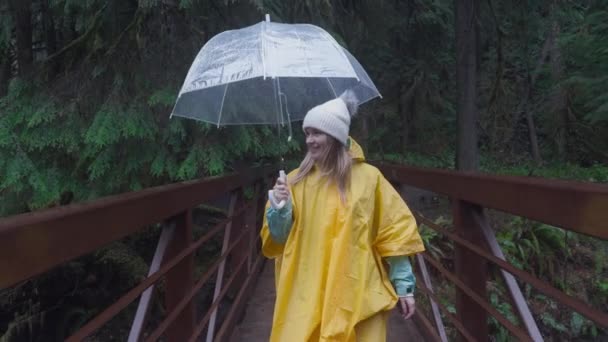 黄色いレインコートの思慮深い女性は 濃い森林に囲まれた橋の上を歩くように自然を熟考しながら 明確な傘を保持しています 雨が降っている フッテージ — ストック動画