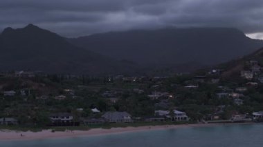 Şiddetli yağmur bulutlarıyla kaplı yüksek dağların ardında altın gün batımı ışığı görülüyor. Oahu Adası, Hawaii 'deki okyanus evleri. Gayrimenkul yatırımı. Lanikai sahilinin yukarısındaki yüksek tepedeki modern sahil kulübeleri.