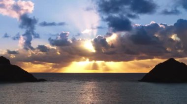 Na Mokulua adalarının üzerinde, Oahu adasının rüzgarlı sahillerinde nefes kesici bir gün doğumu. Parlak altın güneş ışınları okyanusun üzerindeki mavi bulutların arasından parlıyor. Hawaii adalarında dramatik doğa. Oahu macera gezisi
