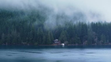 Yoğun sisle kaplı yeşil yağmur ormanlarında gizlenmiş sinematik kır evi. Olimpiyat Ulusal Parkı 'ndaki Crescent Gölü. Sahne Washington doğası 4K USA. Göl manzaralı dağ kulübelerinin hava manzarası.