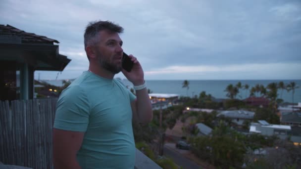 运动微笑的家伙在外面用智能手机说话 有胡子的生意人是自由的长矛手 工作着远程接听电话 在阴天 积极健康的男子站在有热带岛屿景观的阳台上 — 图库视频影像