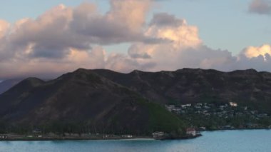 Güneş Hawaii adasının üzerinde bulutları aydınlattı. Yaz tatili geçmişi. 4K Oahu turizmi. Lanikai sahilinin yukarısındaki yeşil tepeye yuva yapmış tatil evleri. Şafak vakti tropikal doğa manzarası üzerinde hava manzarası