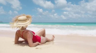 Mavi gökyüzünde güneşlenen bir kadın. Yaz turizmi Hawaii Oahu gezisi. Yan görünümü rahat, ince vücutlu, tropikal plaj arka planında kırmızı seksi mayo giymiş bir manken. İnsanlar cennet adasına seyahat eder.