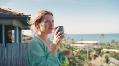 Hawaii adasında tropikal tatilin tadını çıkaran zarif bir kadın. Boş zamanların yaşam tarzı. Gülümseyen kadın plaj köşkünden okyanus manzarasını görmezden geliyor. Balkonda rahat keten gömlekli genç bir kadın ve bir fincan çay.